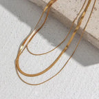 Gold Plated Three-Layer Snake Chain Necklace displayed on a textured surface with sunlight highlighting its elegant, smooth, and sleek design in warm gold tones.