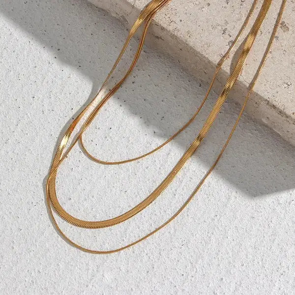 Gold Plated Three-Layer Snake Chain Necklace displayed on a textured surface with sunlight highlighting its elegant, smooth, and sleek design in warm gold tones.