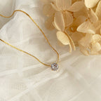 Old-Tone Solitaire Pendant Necklace featuring a delicate gold chain and a single sparkling gemstone, styled on soft white fabric with beige dried flowers for an elegant look.