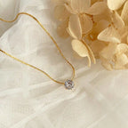 Old-Tone Solitaire Pendant Necklace featuring a delicate gold chain and a single sparkling gemstone, styled on soft white fabric with beige dried flowers for an elegant look.