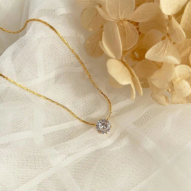 Old-Tone Solitaire Pendant Necklace featuring a delicate gold chain and a single sparkling gemstone, styled on soft white fabric with beige dried flowers for an elegant look.