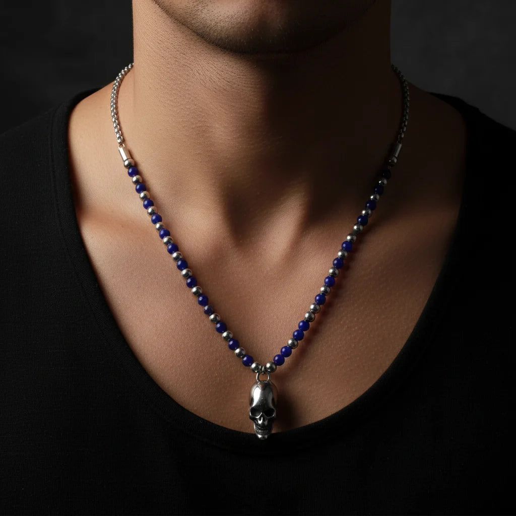 A close-up of a man's neck and chest wearing a beaded necklace with a silver skull pendant. The necklace features deep blue and metallic silver beads on a dark chain, styled against a black shirt.
