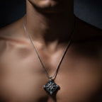 A close-up of a man's neck and chest wearing a silver chain necklace with a diamond-shaped pendant featuring a central floral or snowflake-like motif and small clear stones at the corners.