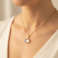 A close-up of a model wearing a delicate gold chain necklace with a round bezel-set iridescent moonstone pendant, styled with a soft cream-colored top.