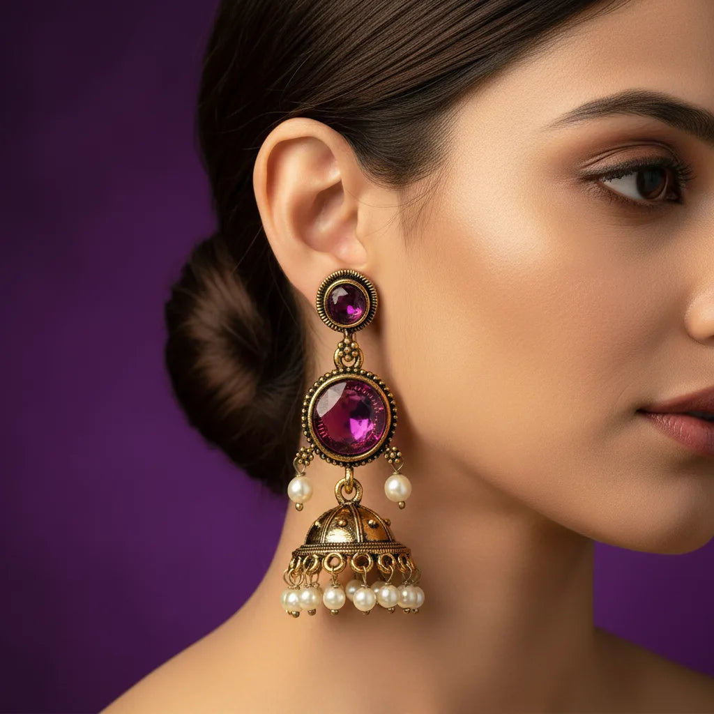 A close-up of a woman's ear wearing an ornate gold-toned dangle earring with a large purple gemstone and small pearl drops, set against a solid purple background.