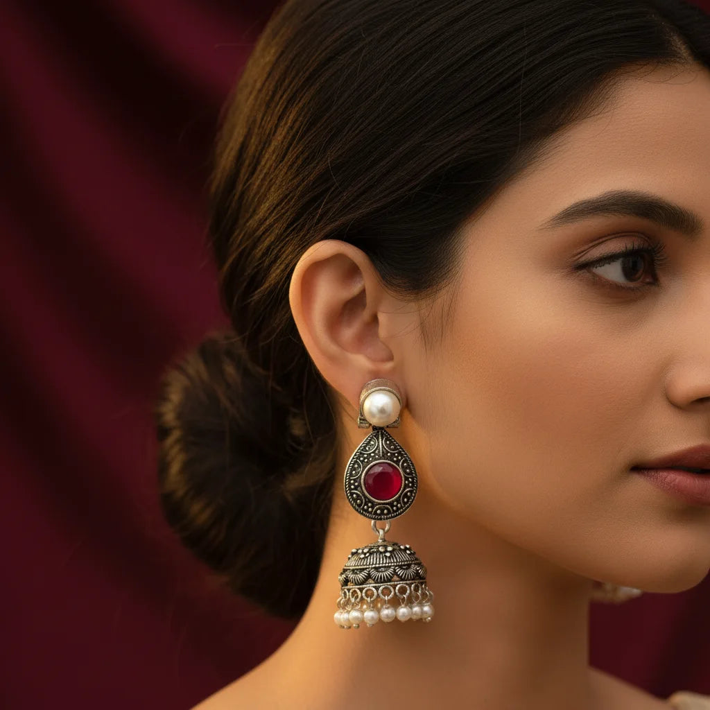 A close-up of a woman wearing an ornate silver-toned jhumka earring with a red teardrop-shaped stone and a white pearl at the top, set against a dark burgundy background.