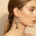 A close-up side profile of a woman wearing a dangling gold earring with clear teardrop-shaped stones and a small clear stone cluster at the top, against a soft beige background.