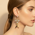 A close-up side profile of a woman wearing a dangling gold earring with clear teardrop-shaped stones and a small clear stone cluster at the top, against a soft beige background.