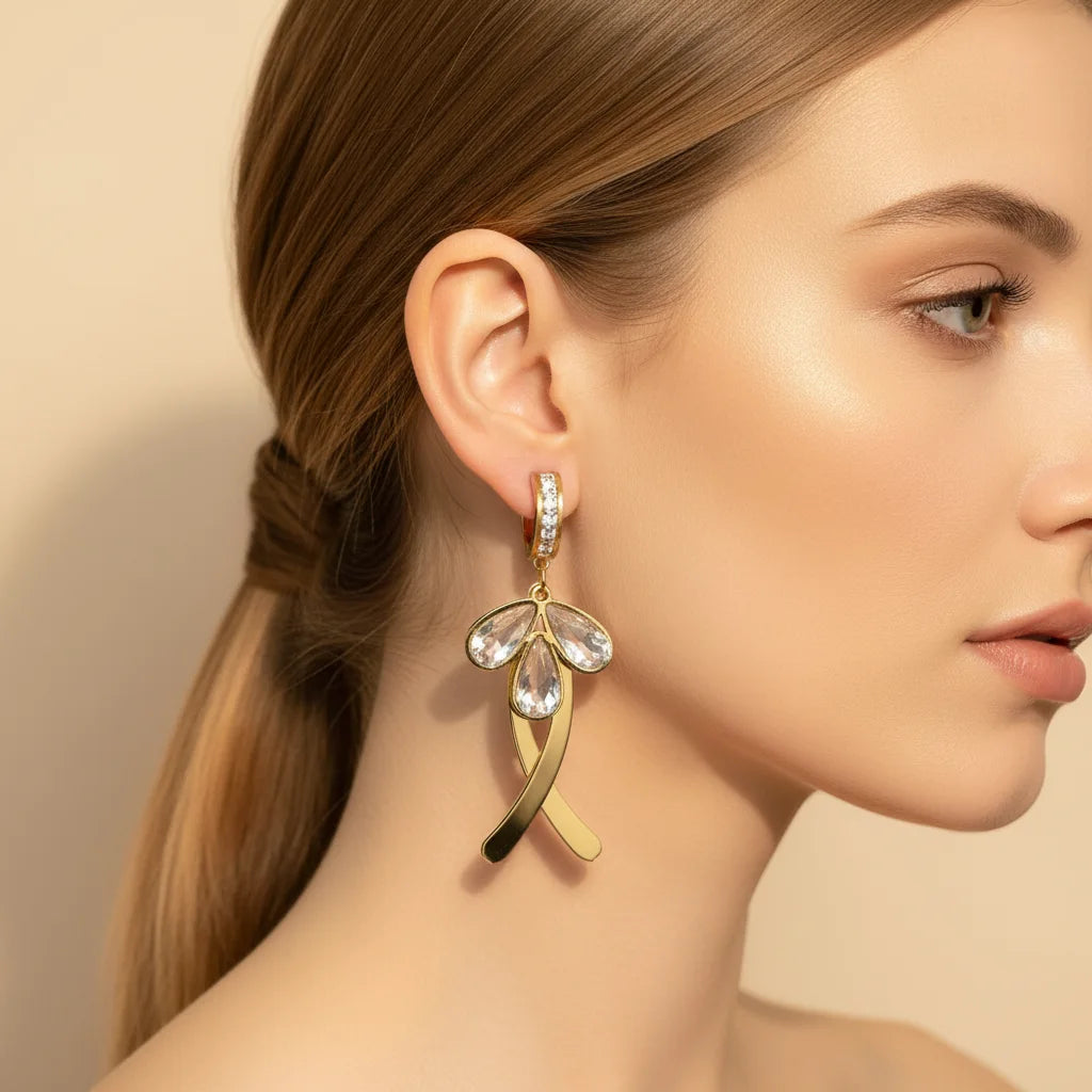 A close-up side profile of a woman wearing a dangling gold earring with clear teardrop-shaped stones and a small clear stone cluster at the top, against a soft beige background.