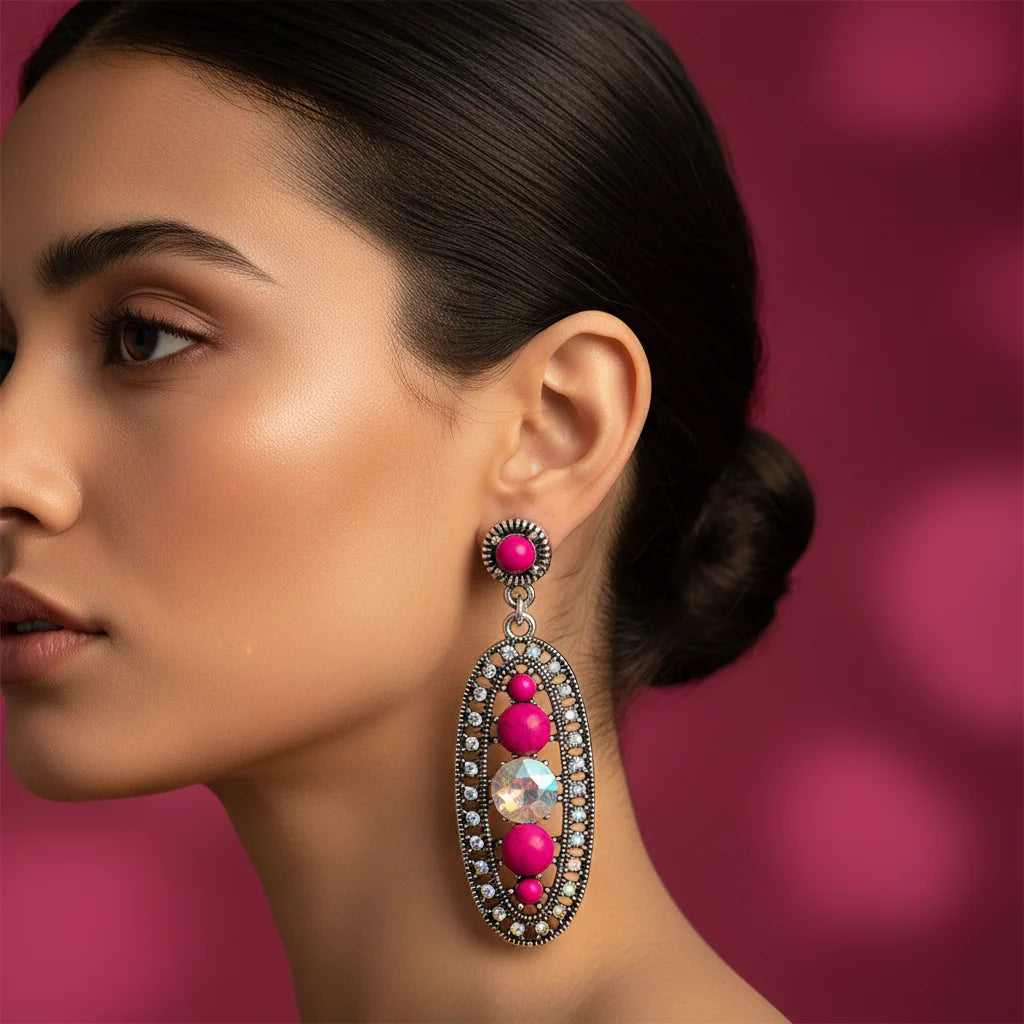 A close-up side profile of a woman wearing a dramatic drop earring. The earring features bright pink gemstone beads set in silver-toned metal with small clear stones, set against a soft pink bokeh background.