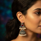 A close-up side profile of a woman wearing a large silver-toned jhumka earring with a red teardrop stone, small green and blue stones, and dangling pearl beads against a dark blurred background.