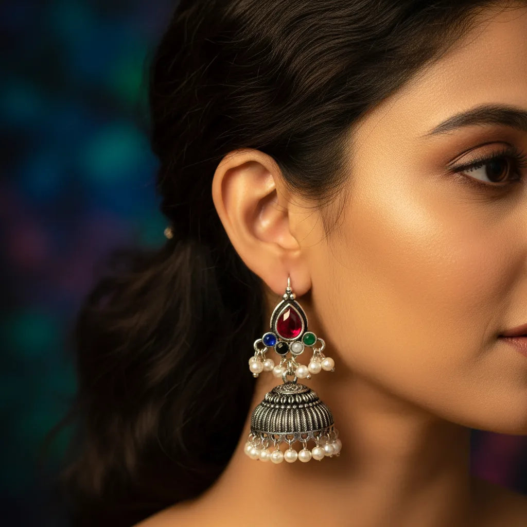 A close-up side profile of a woman wearing a large silver-toned jhumka earring with a red teardrop stone, small green and blue stones, and dangling pearl beads against a dark blurred background.