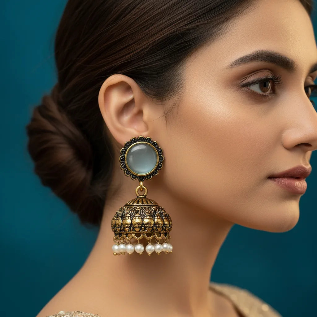 A close-up side profile of a woman wearing a large traditional Indian jhumka earring. The earring features a round pale blue stone in a black and gold frame, hanging from an ornate gold-toned bell-shaped dangle decorated with small white pearls, set against a dark teal background.