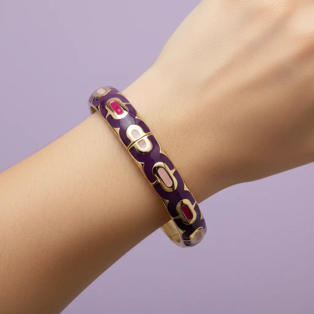A gold-plated bangle bracelet with a deep purple enamel background and small red oval accents, shown worn on a person's wrist against a solid lavender background.