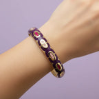 A gold-plated bangle bracelet with a deep purple enamel background and small red oval accents, shown worn on a person's wrist against a solid lavender background.