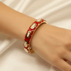 A gold-plated bangle bracelet with a red enamel background and repeating cream-colored oval links, shown worn on a person's wrist against a soft white fabric backdrop.