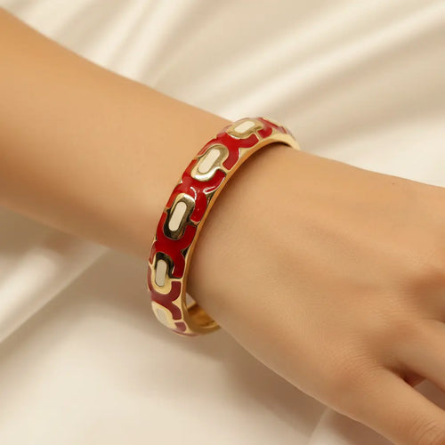 A gold-plated bangle bracelet with a red enamel background and repeating cream-colored oval links, shown worn on a person's wrist against a soft white fabric backdrop.