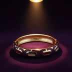 A gold-plated bangle bracelet with purple and white enamel cutouts, displayed on a dark purple surface under a spotlight.