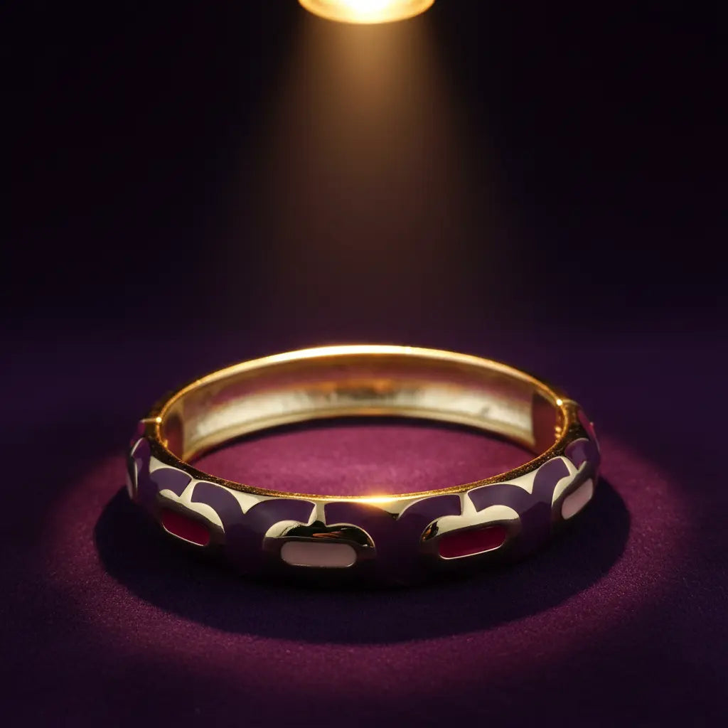 A gold-plated bangle bracelet with purple and white enamel cutouts, displayed on a dark purple surface under a spotlight.