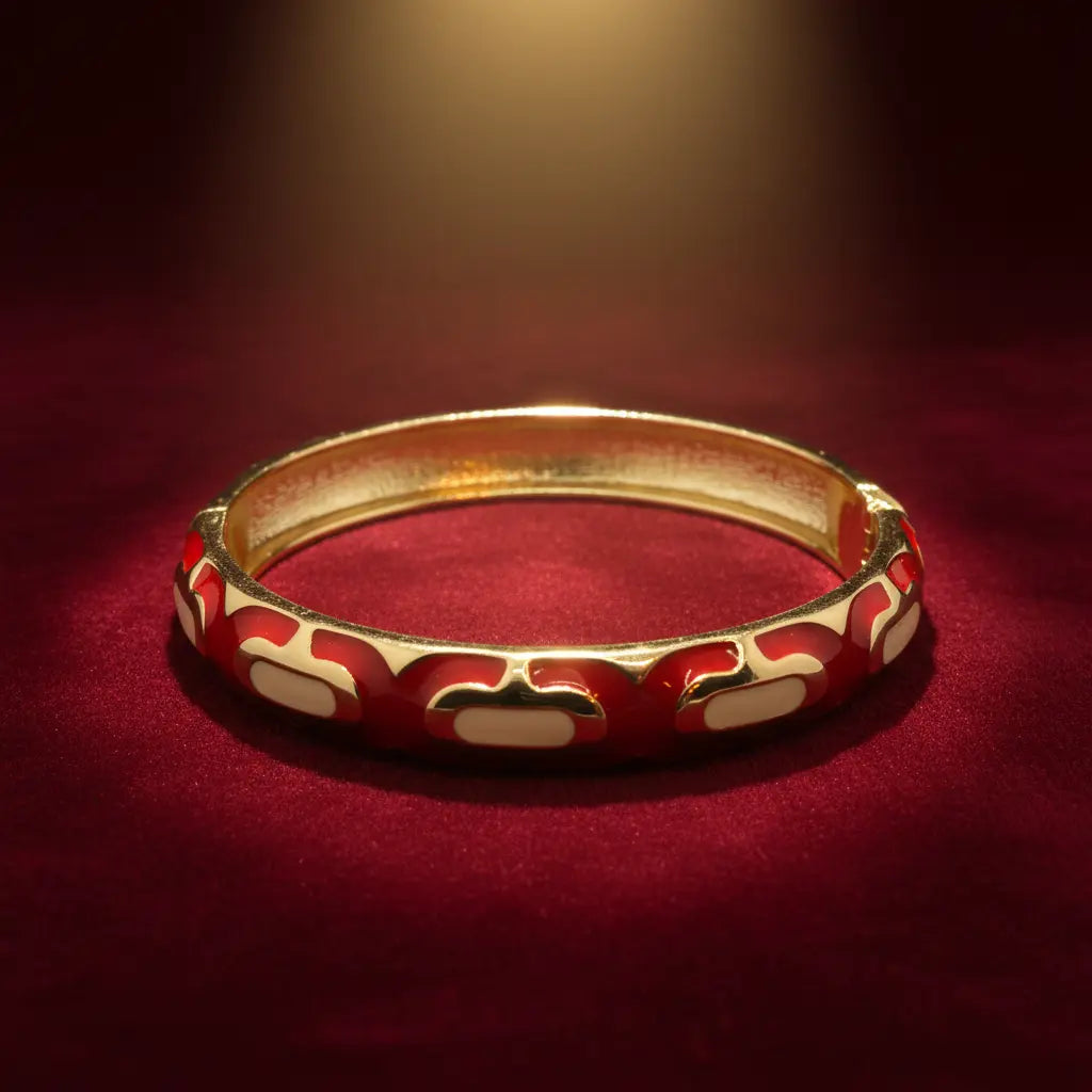 A gold-plated bangle bracelet with red and cream enamel patterns, displayed on a dark red velvet surface under warm spotlighting.