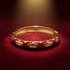 A gold-plated bangle bracelet with red and cream enamel patterns, displayed on a dark red velvet surface under warm spotlighting.