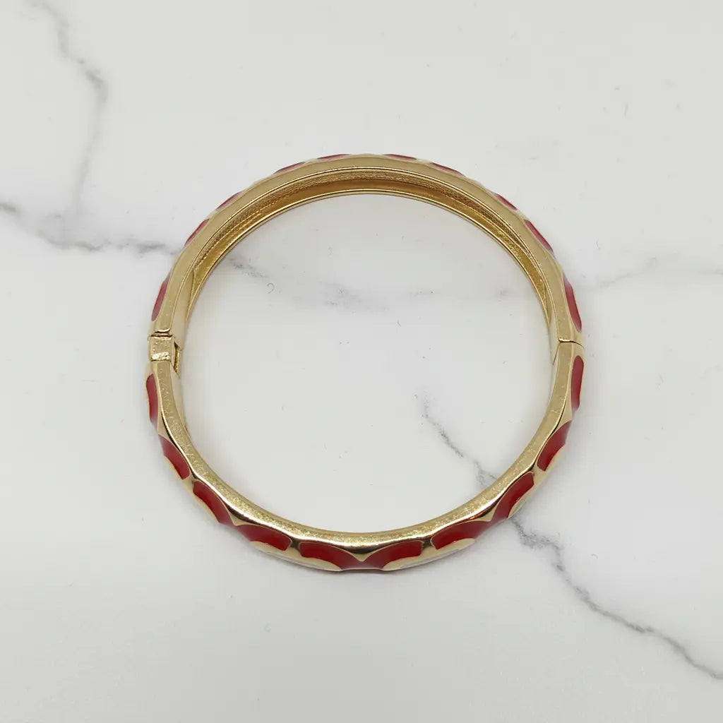 A gold-plated bangle bracelet with red enamel inlay, displayed on a white marble surface.