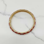 A gold-plated bangle bracelet with red enamel inlay, displayed on a white marble surface.