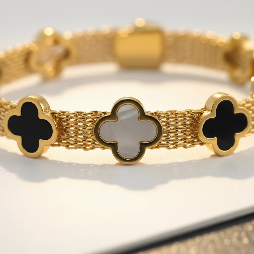 A gold-tone mesh bracelet with four clover-shaped links, featuring black inlay on two ends and a central white inlay clover, photographed close-up on a light surface.