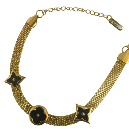 A gold-tone mesh chain necklace with three floral clover motifs, two featuring black enamel inlays and one solid gold-tone clover, displayed on a light off-white background.