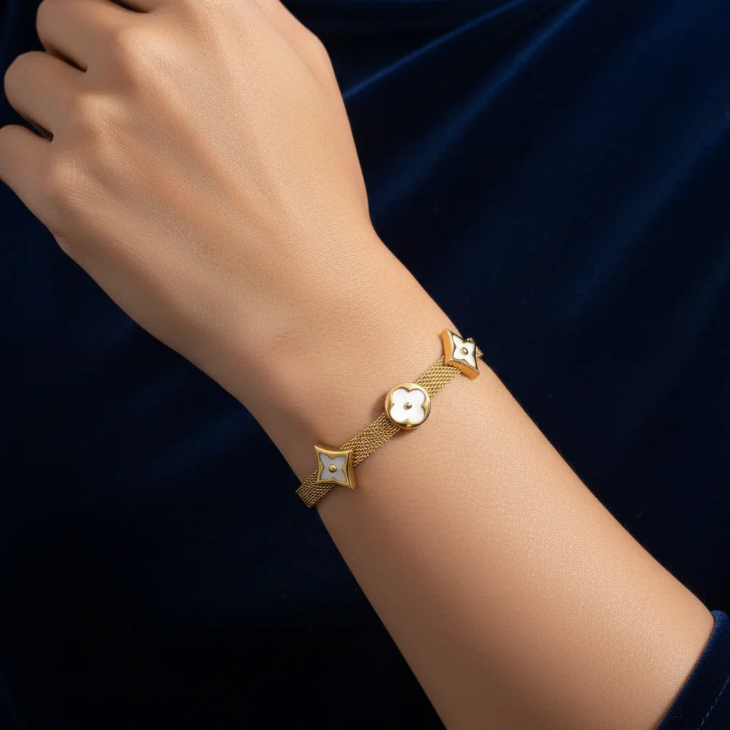 A gold bracelet with three decorative charms (a central clover and two star-like shapes with inlay details) worn on a person's wrist against a dark navy blue velvet background.