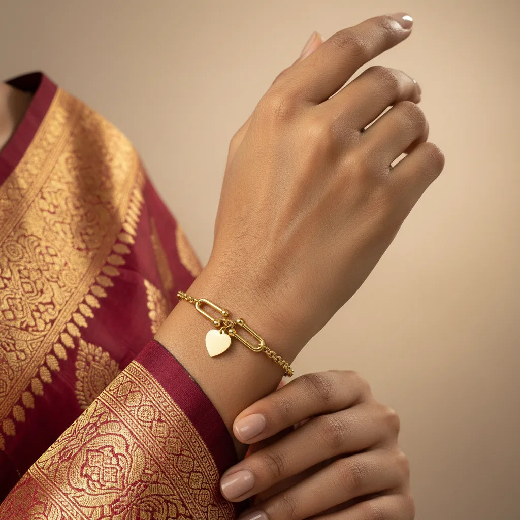 A gold chain bracelet with a heart-shaped charm and elongated link details, shown worn on a woman's wrist. The person is wearing a deep red and gold patterned traditional garment, set against a soft beige background.