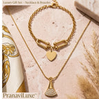 A gold necklace and bracelet gift set by PranaviLuxe, featuring a delicate gold chain necklace with a fan-shaped pendant studded with small clear crystals, and a matching gold chain bracelet with a heart charm and elongated link detail, styled on a pink marble surface with a patterned scarf and dried grass.