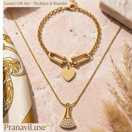 A gold necklace and bracelet gift set by PranaviLuxe, featuring a delicate gold chain necklace with a fan-shaped pendant studded with small clear crystals, and a matching gold chain bracelet with a heart charm and elongated link detail, styled on a pink marble surface with a patterned scarf and dried grass.
