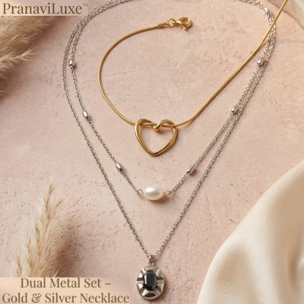 A layered double necklace set by PranaviLuxe, featuring a gold chain with a heart-shaped pendant and a silver chain with a white pearl and a silver bezel-set stone pendant, displayed on a textured beige surface with dried grass and satin fabric.