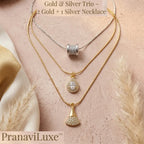 A layered necklace set displayed on a soft pink textured surface with decorative dried grass and fabric. The set includes a silver chain with a silver bead pendant, a gold chain with a gold teardrop pendant set with clear stones, and a gold chain with a gold fan-shaped pendant set with clear stones.