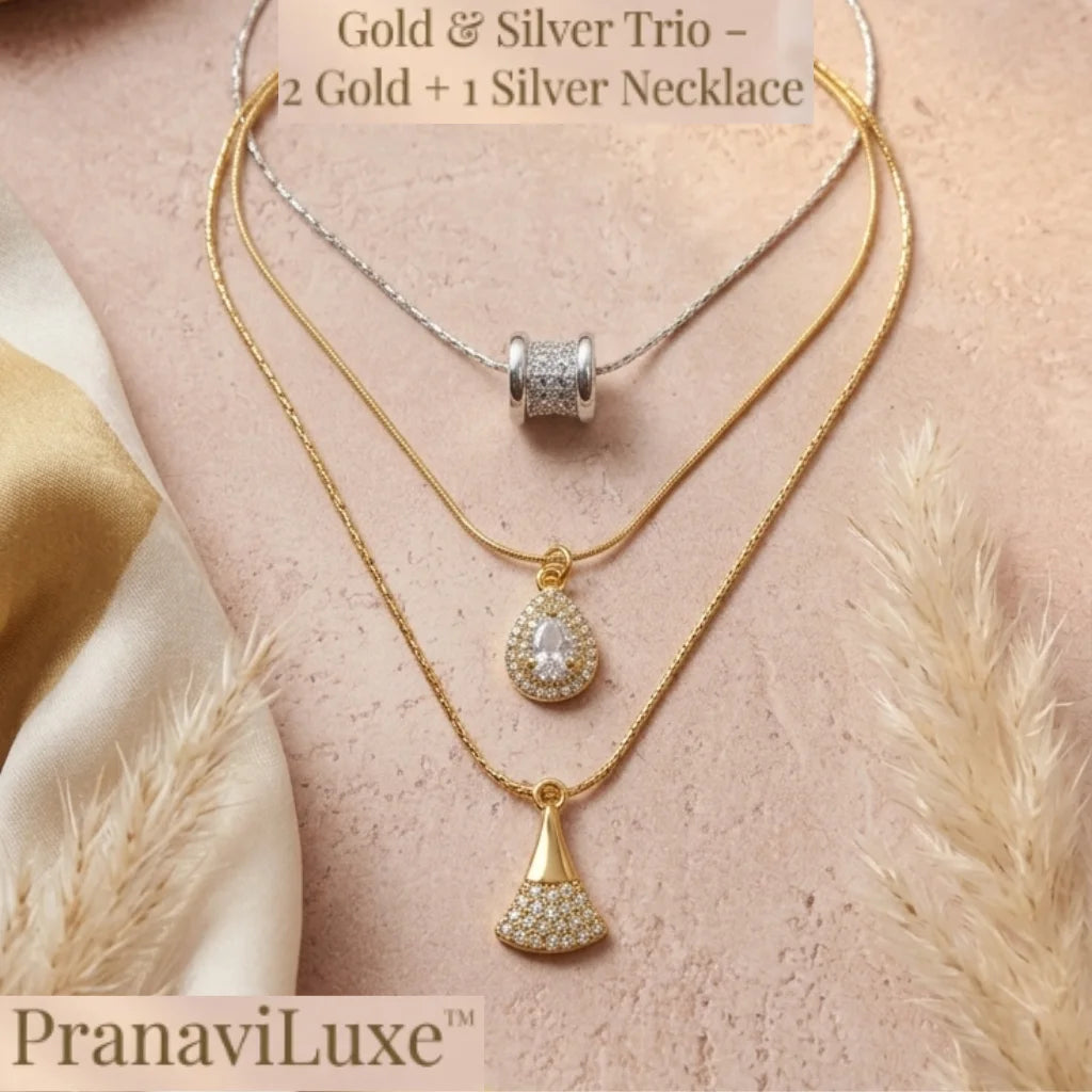 A layered necklace set displayed on a soft pink textured surface with decorative dried grass and fabric. The set includes a silver chain with a silver bead pendant, a gold chain with a gold teardrop pendant set with clear stones, and a gold chain with a gold fan-shaped pendant set with clear stones.