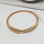 A rose gold bangle bracelet with engraved Roman numerals and a row of clear crystals, resting on a white marble surface.