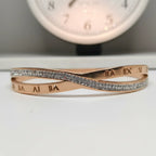 A rose gold infinity-style bangle bracelet with pavé-set clear stones across the center and Roman numeral engravings on the inner band, displayed on a white surface with a blurred clock in the background.