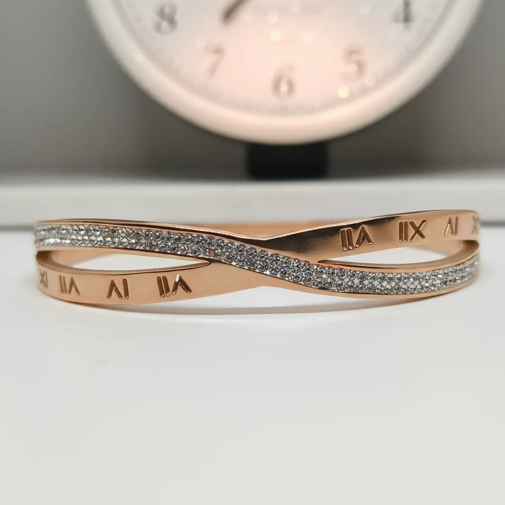 A rose gold infinity-style bangle bracelet with pavé-set clear stones across the center and Roman numeral engravings on the inner band, displayed on a white surface with a blurred clock in the background.