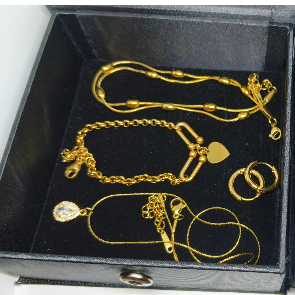 A set of gold jewelry pieces, including a chain necklace, bracelet, rings, and pendant, displayed inside a black presentation box.