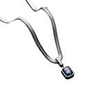 A silver-toned snake chain necklace with a rectangular blue gemstone pendant, displayed on a textured black cylindrical stand against a dark gray background.