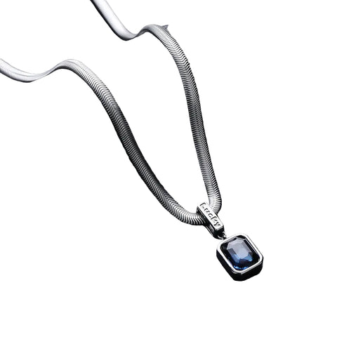 A silver-toned snake chain necklace with a rectangular blue gemstone pendant, displayed on a textured black cylindrical stand against a dark gray background.