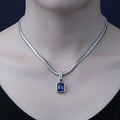 A silver snake chain necklace with a rectangular blue gemstone pendant, displayed on a mannequin neck against a dark background.