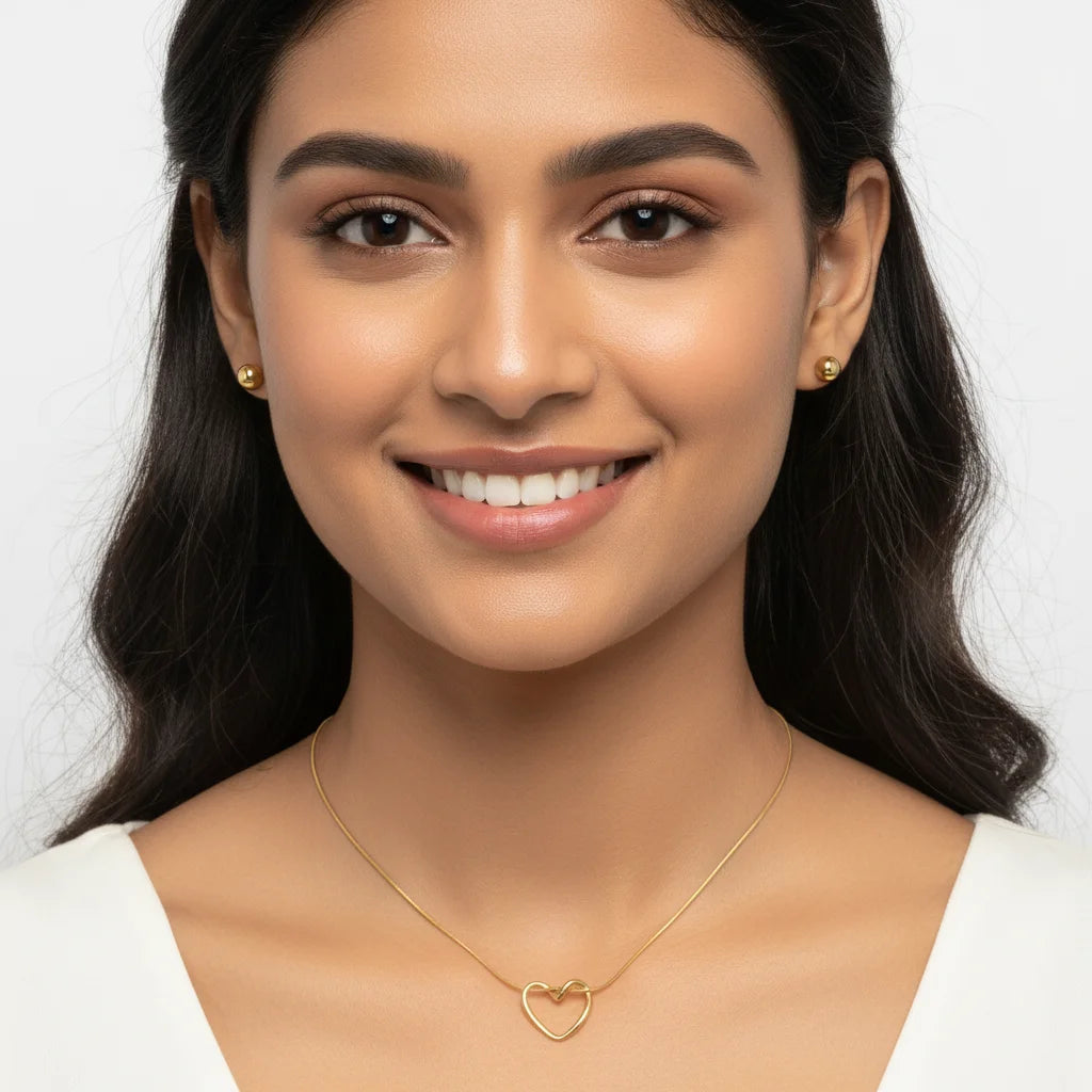 A smiling woman with dark hair wearing a delicate gold heart-link necklace and matching small gold stud earrings against a clean white background.