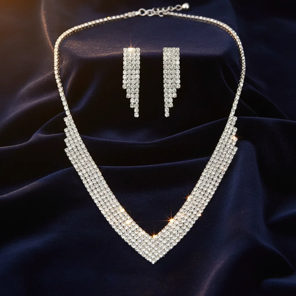 A sparkling silver-tone jewelry set featuring a V-shaped rhinestone necklace and matching drop earrings, displayed on a dark navy blue satin fabric background.