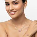 A woman wearing a delicate gold floral necklace with pink and white flower details, matching stud earrings, and a bracelet against a clean white background.