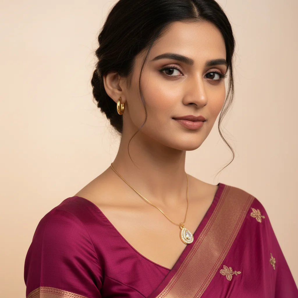 A woman wearing a gold teardrop pendant necklace and matching gold hoop earrings, paired with a magenta saree with gold embroidery, set against a solid beige background.
