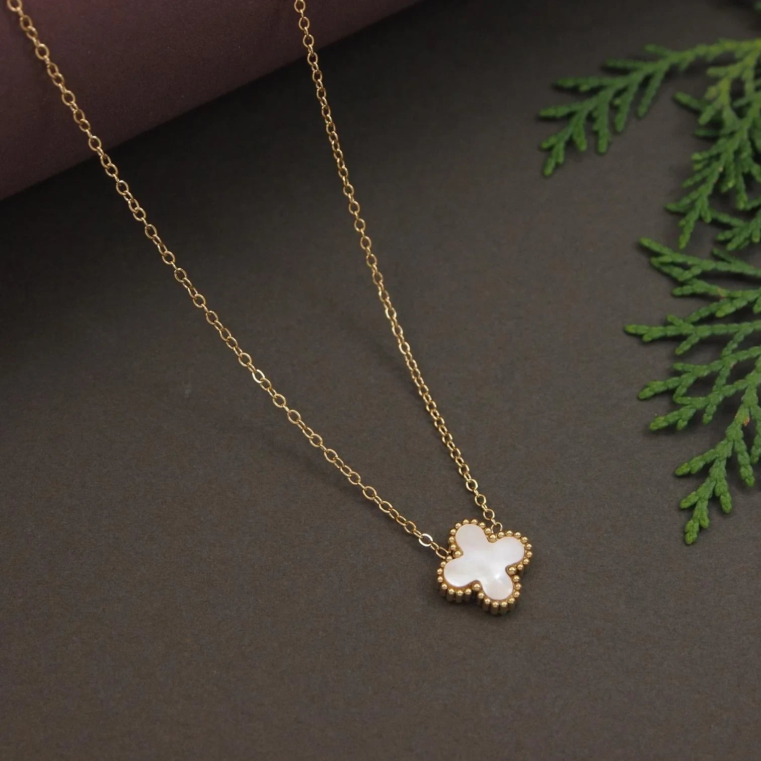 Gold necklace with a clover-shaped pendant on a dark surface with green leaves.