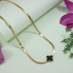 Delicate gold-toned Lucky Clover Snake Chain Necklace with a black four-leaf clover pendant, styled against a soft white background with floral accents.