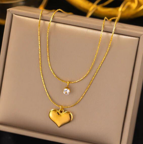 Crystal Heart Double Layered Necklace featuring a delicate gold chain with a small clear crystal pendant on the upper layer and a polished gold heart pendant on the lower layer, elegant and modern jewelry.